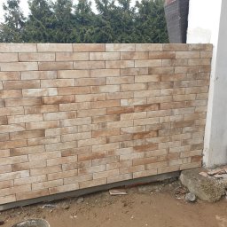 Sławomir Michalski - Ściana wykończona beżowo-brązową cegłą klinkierową, widoczne zielone drzewa w tle. Fragment ściany budynku z niedokończonym wykończeniem po prawej stronie.