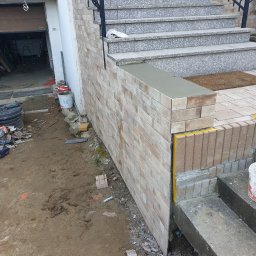 Sławomir Michalski - Murowana ściana z cegły klinkierowej przy schodach wejściowych, z widocznymi elementami wykończeniowymi i narzędziami budowlanymi w tle. Detal prac murarskich.