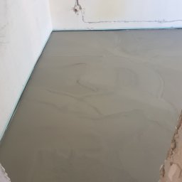 Sławomir Michalski - Świeżo wylana, mokra wylewka podłogowa w rogu pomieszczenia, widoczne ślady po rozprowadzaniu masy. Przy ścianach listwa dylatacyjna.