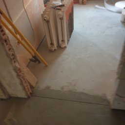 Sławomir Michalski - Wyrównana, szara posadzka w trakcie prac remontowych. W tle grzejnik żeberkowy, narzędzia i fragment ściany z widoczną warstwą izolacyjną. Ujęcie z niskiej perspektywy.