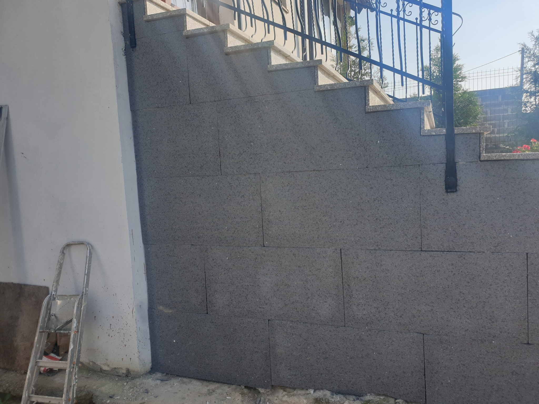 Szare, prostokątne płyty elewacyjne ułożone na ścianie przy schodach zewnętrznych z metalową barierką. Drabina oparta o ścianę z lewej strony.