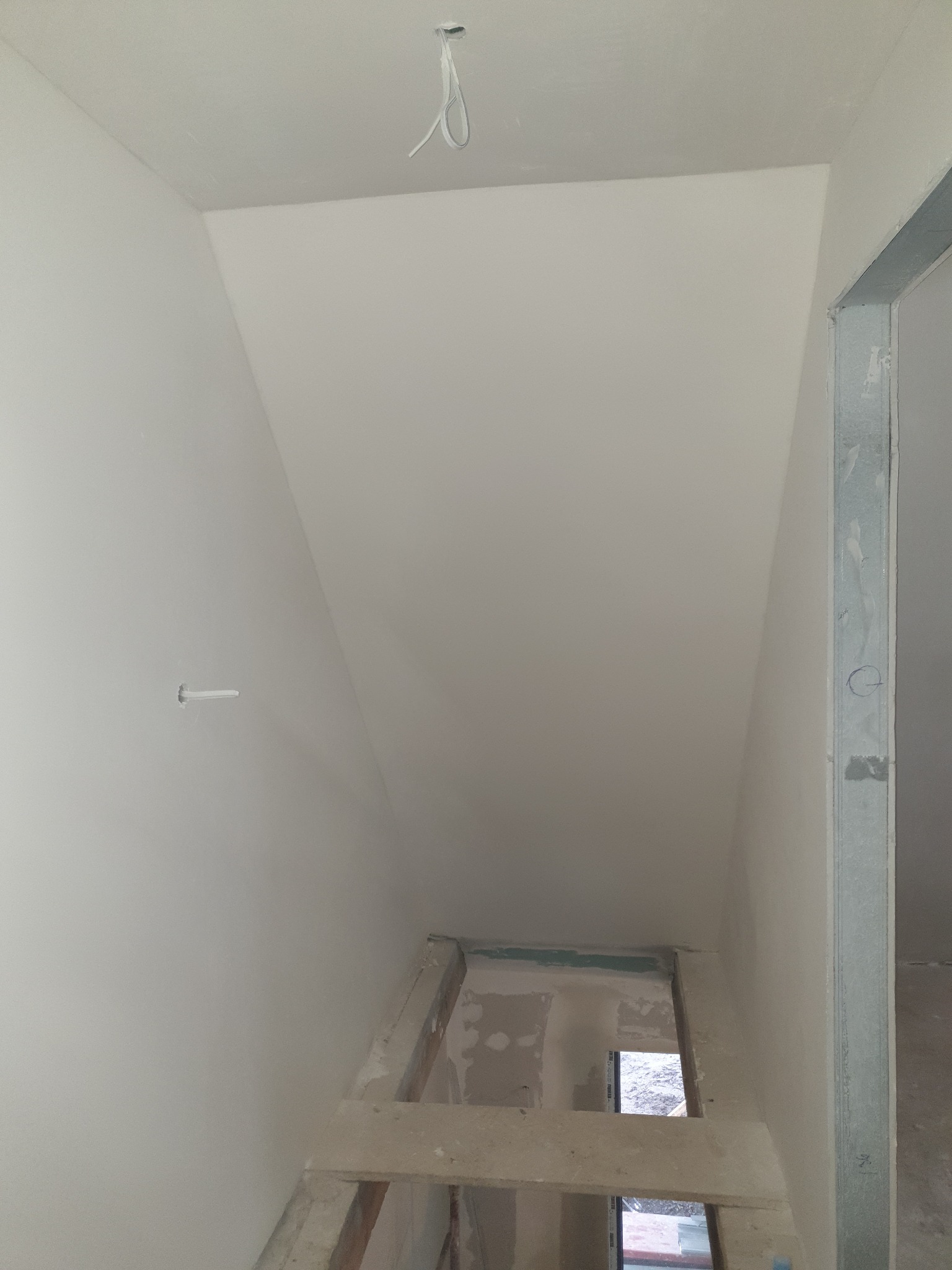 Wnętrze poddasza po remoncie, ściany i sufit wykończone na gładko, widoczne elementy konstrukcyjne i instalacja elektryczna. Metalowa rama drzwiowa.