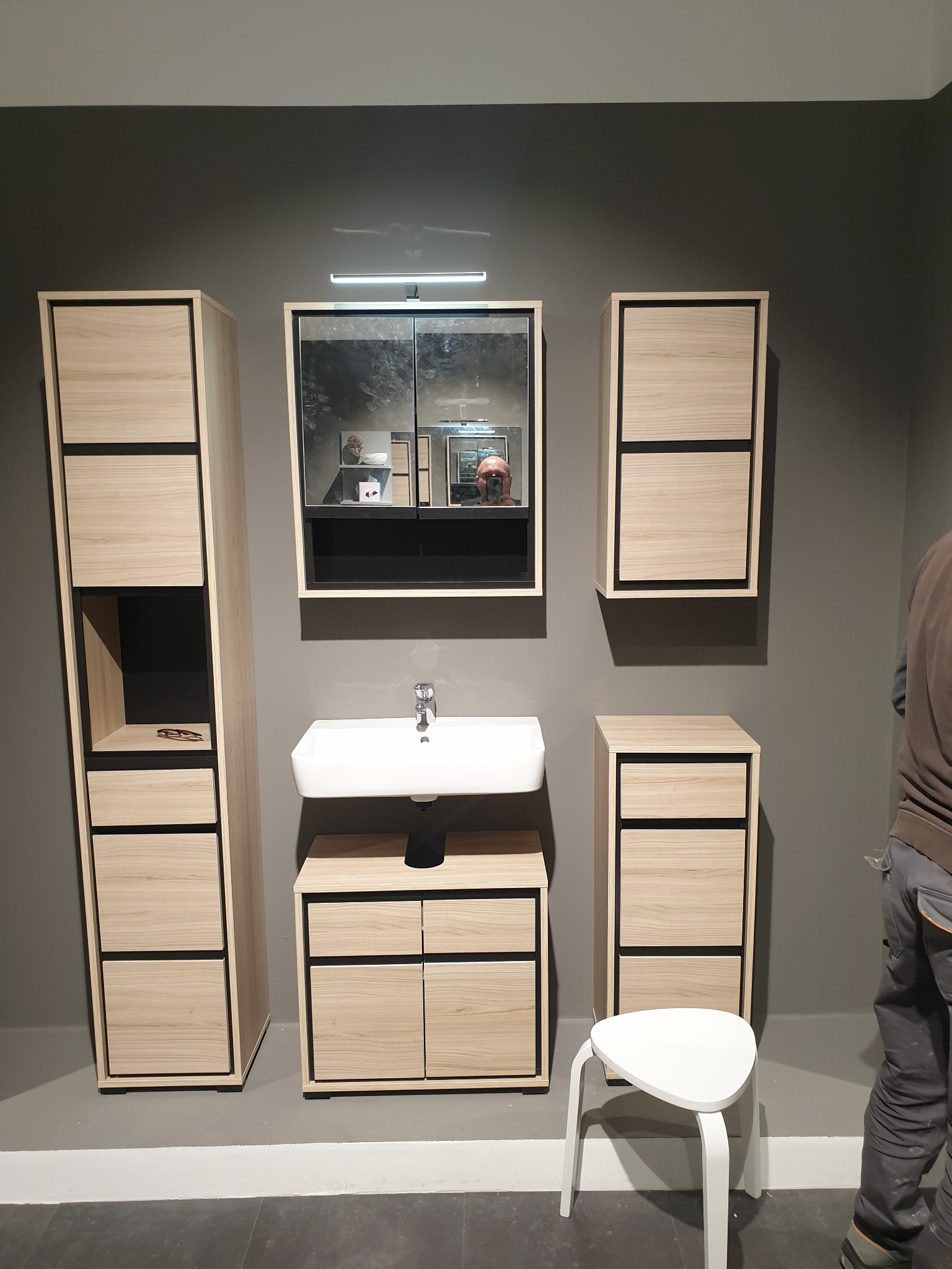 Nowoczesna aranżacja łazienki z drewnianymi szafkami z czarnymi akcentami, umywalką i lustrem z oświetleniem. Widoczny fragment osoby w odbiciu lustra.