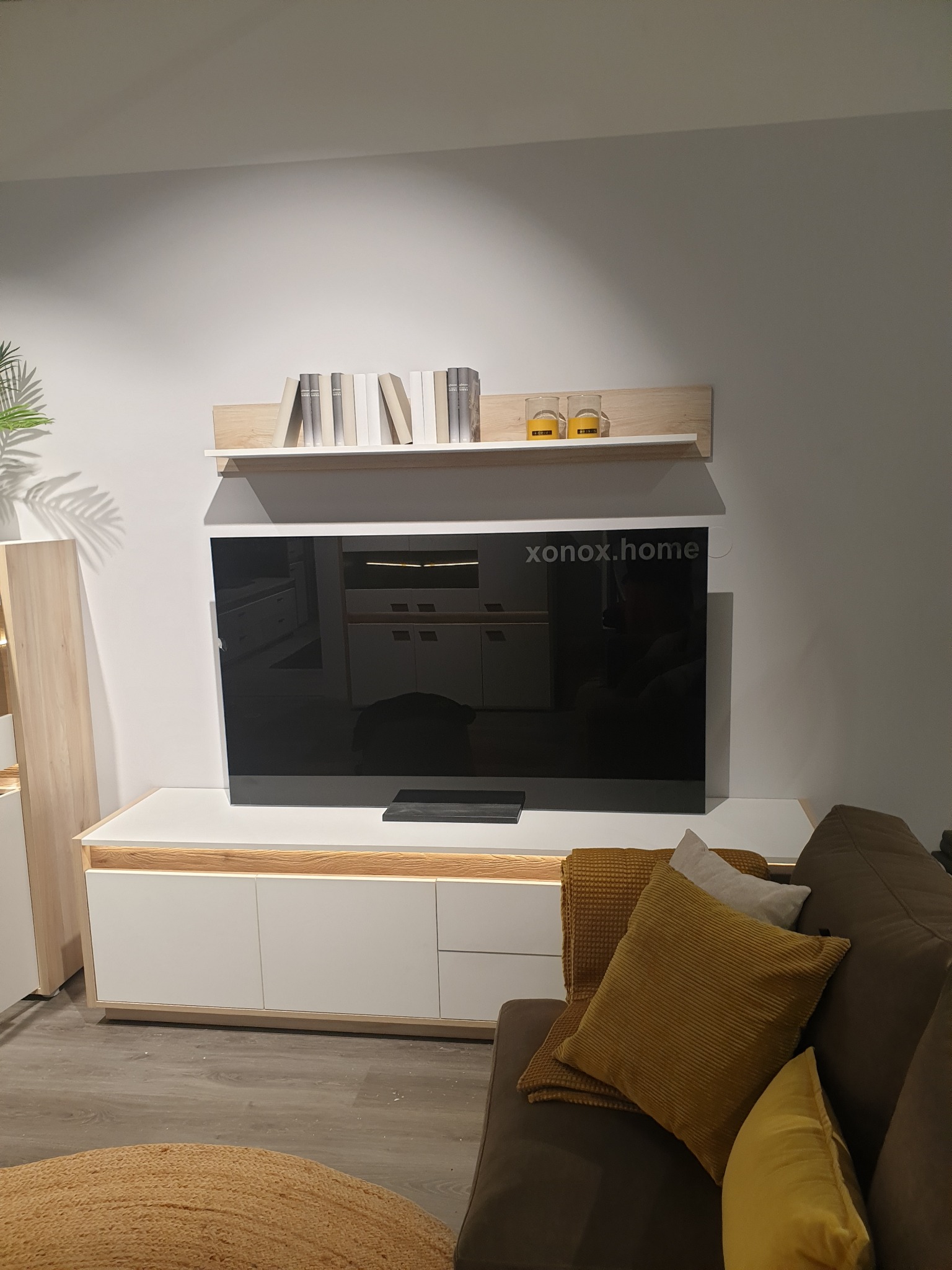 Nowoczesny salon z telewizorem na szafce RTV, półką z książkami i dekoracjami. Minimalistyczny design, ciepłe oświetlenie, brązowa sofa z poduszkami.