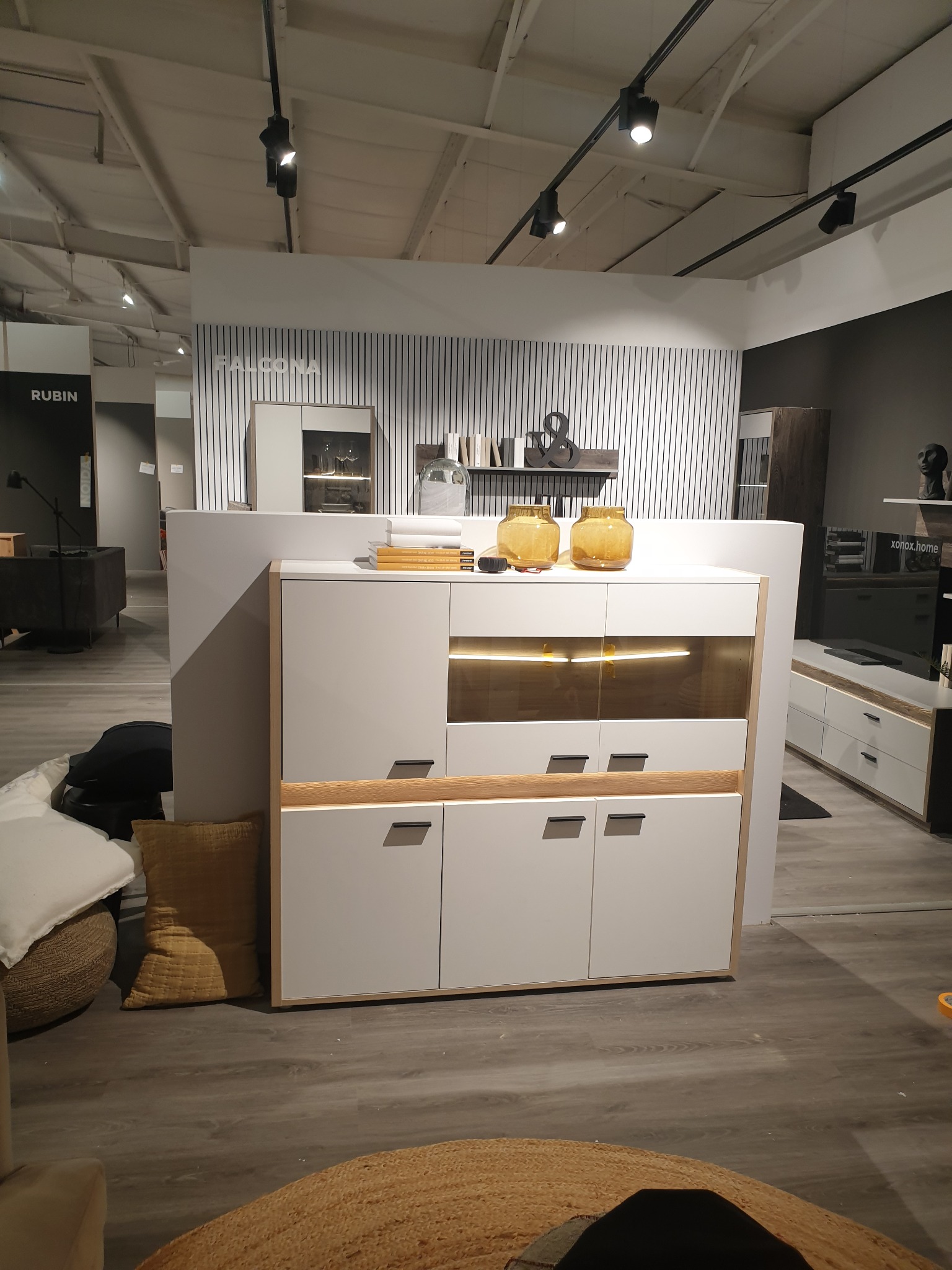 Nowoczesna biała komoda z drewnianymi akcentami, oświetlona od wewnątrz, na tle minimalistycznego wnętrza z dekoracyjną ścianą w paski i elementami wyposażenia.