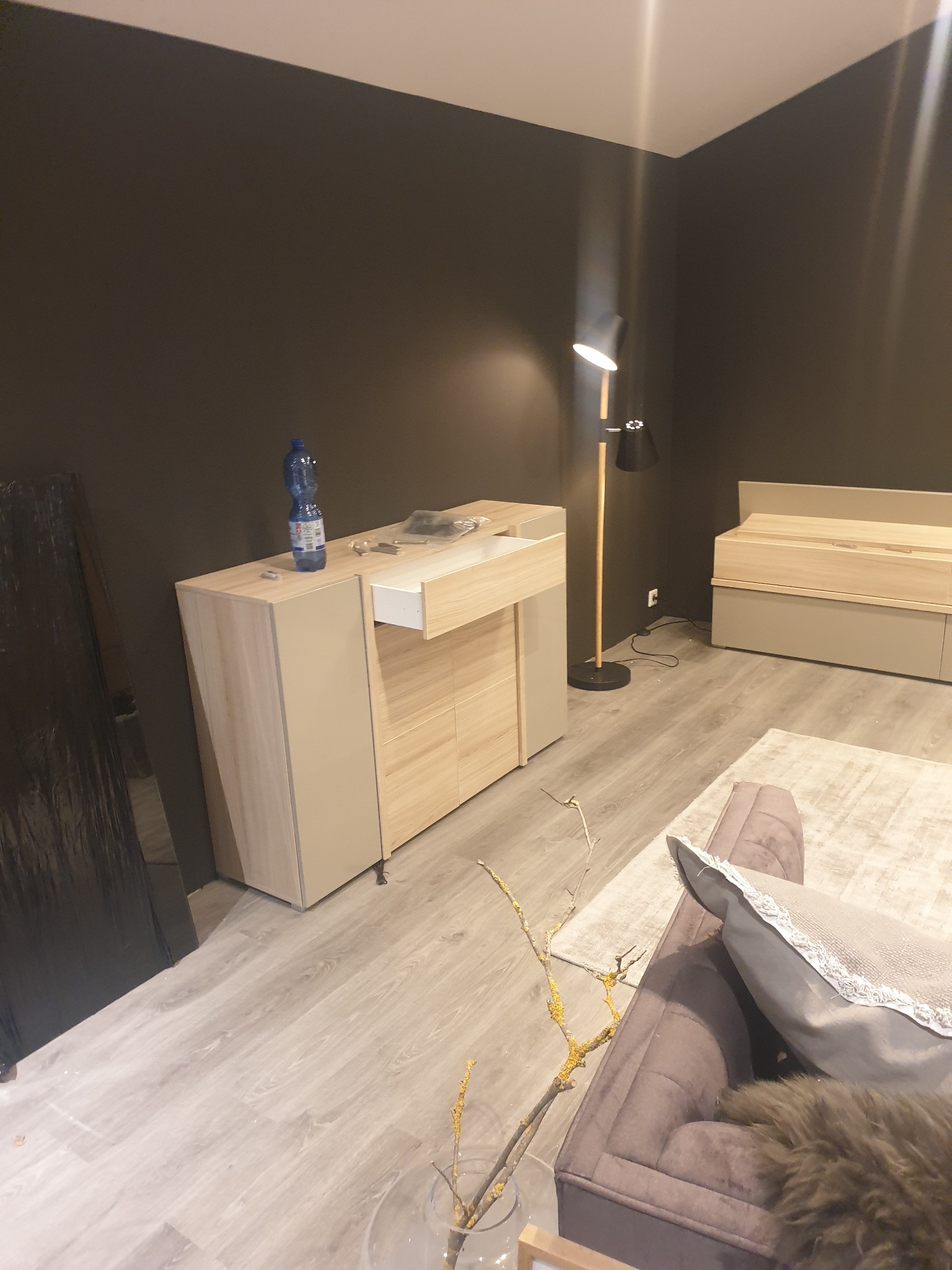 Ściana pomalowana na ciemny kolor w pokoju z drewnianą komodą z wysuniętą szufladą, lampą i sofą. Widoczna butelka wody na komodzie.