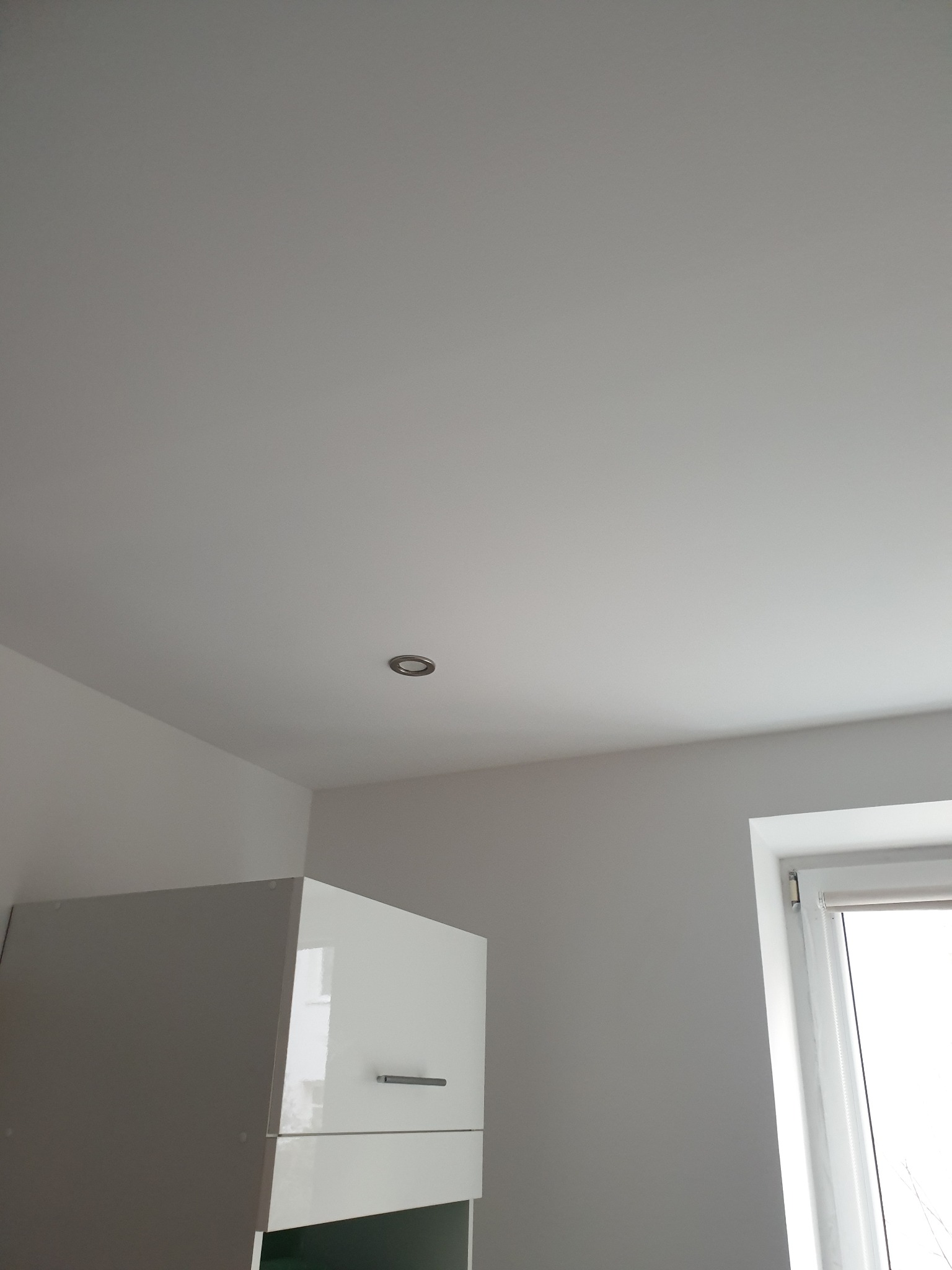 Biały, gładki sufit z wbudowanym punktowym oświetleniem, fragment szafki kuchennej i okno z roletą w tle. Minimalistyczne wnętrze po remoncie.