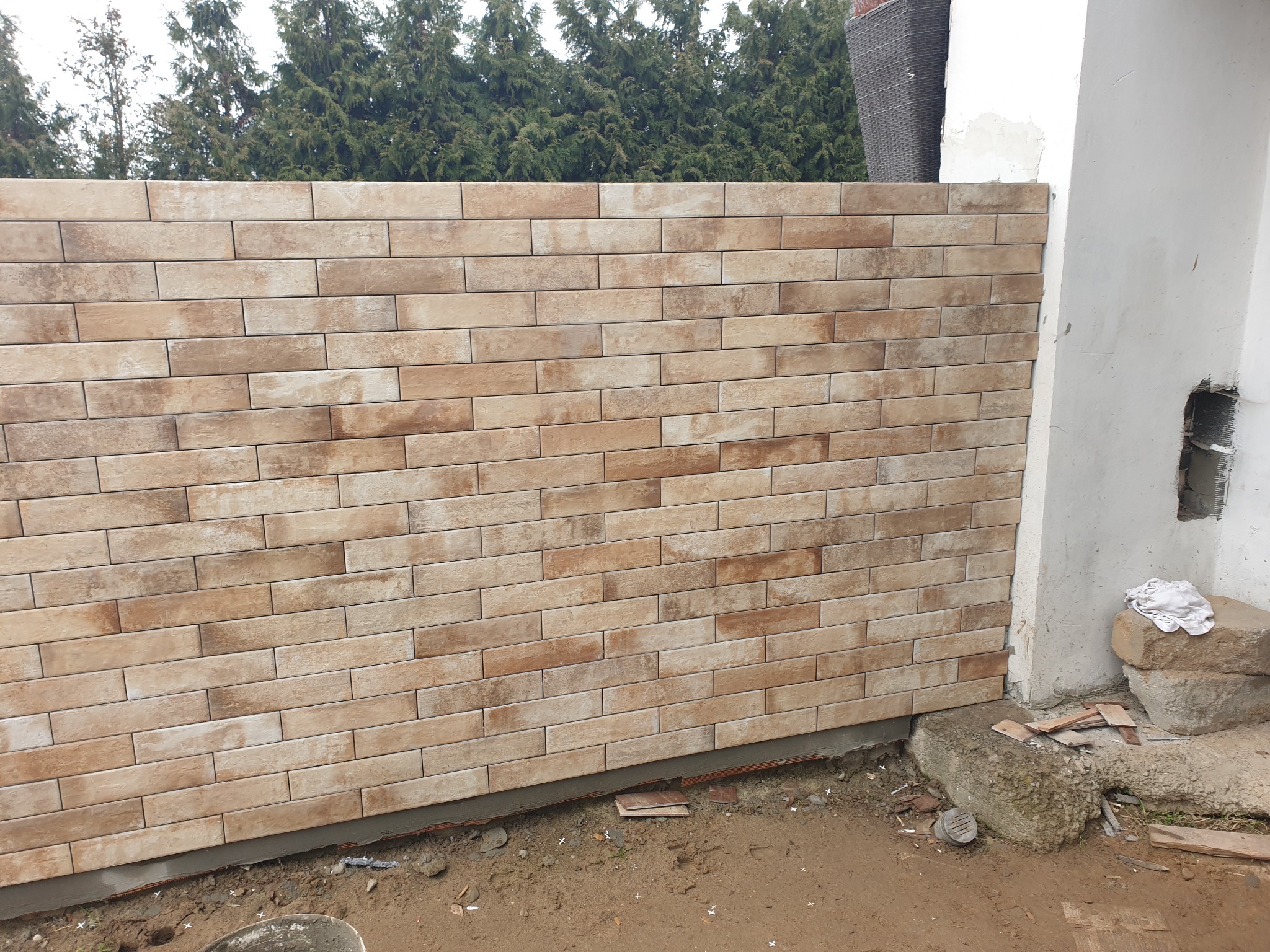 Ściana wykończona beżowo-brązową cegłą klinkierową, widoczne zielone drzewa w tle. Fragment ściany budynku z niedokończonym wykończeniem po prawej stronie.
