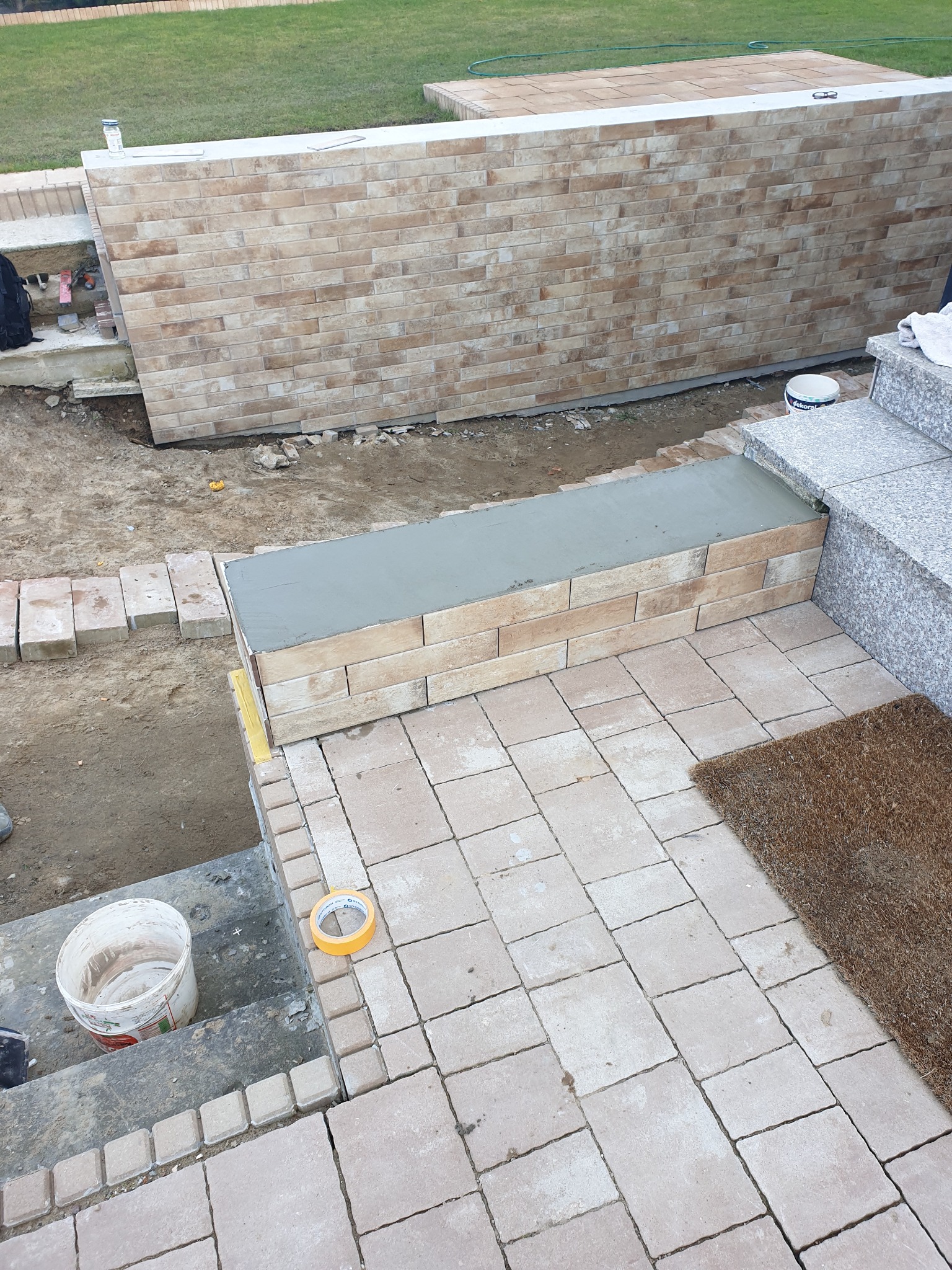 Układanie kostki brukowej na tarasie z elementami murowanymi. Widoczny etap prac z użyciem zaprawy i taśmy malarskiej. Tło: trawnik.