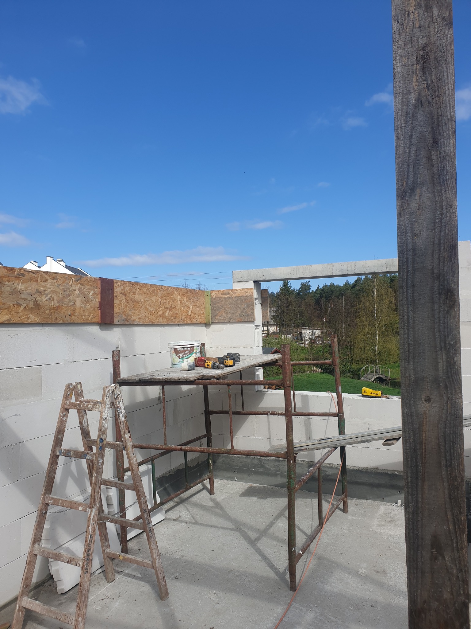 Budowa domu z białych bloczków: ściany w trakcie wznoszenia, rusztowanie z narzędziami, drabina malarska. Widok na zewnątrz przez otwór okienny, błękitne niebo.