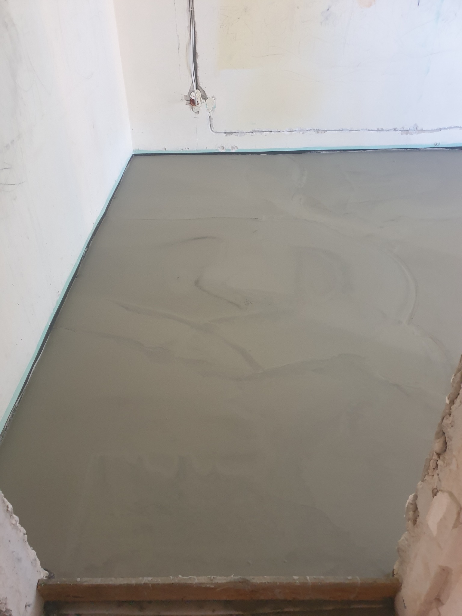 Świeżo wylana, mokra wylewka podłogowa w rogu pomieszczenia, widoczne ślady po rozprowadzaniu masy. Przy ścianach listwa dylatacyjna.