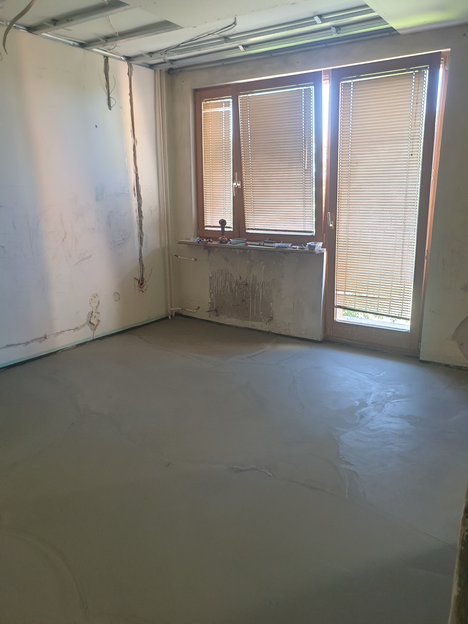 Wyrównana, świeża wylewka podłogowa w pokoju z oknem i grzejnikiem, przygotowana do dalszych prac wykończeniowych w Elblągu. Widoczne ślady po instalacjach.