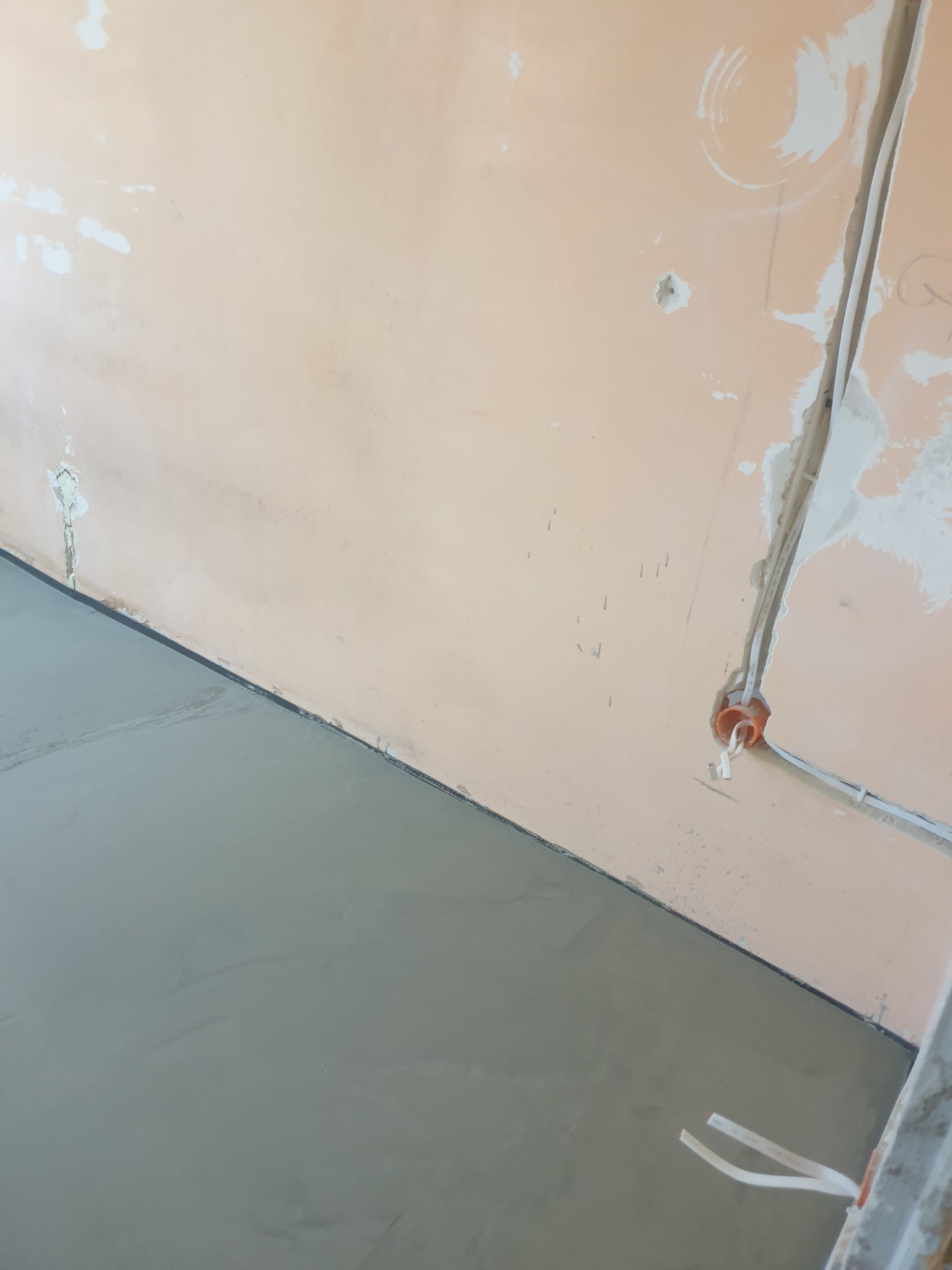Świeżo wylana, szara wylewka podłogowa w pomieszczeniu z przygotowaną instalacją elektryczną w ścianie. Widoczne ślady po pracach remontowych na ścianie.