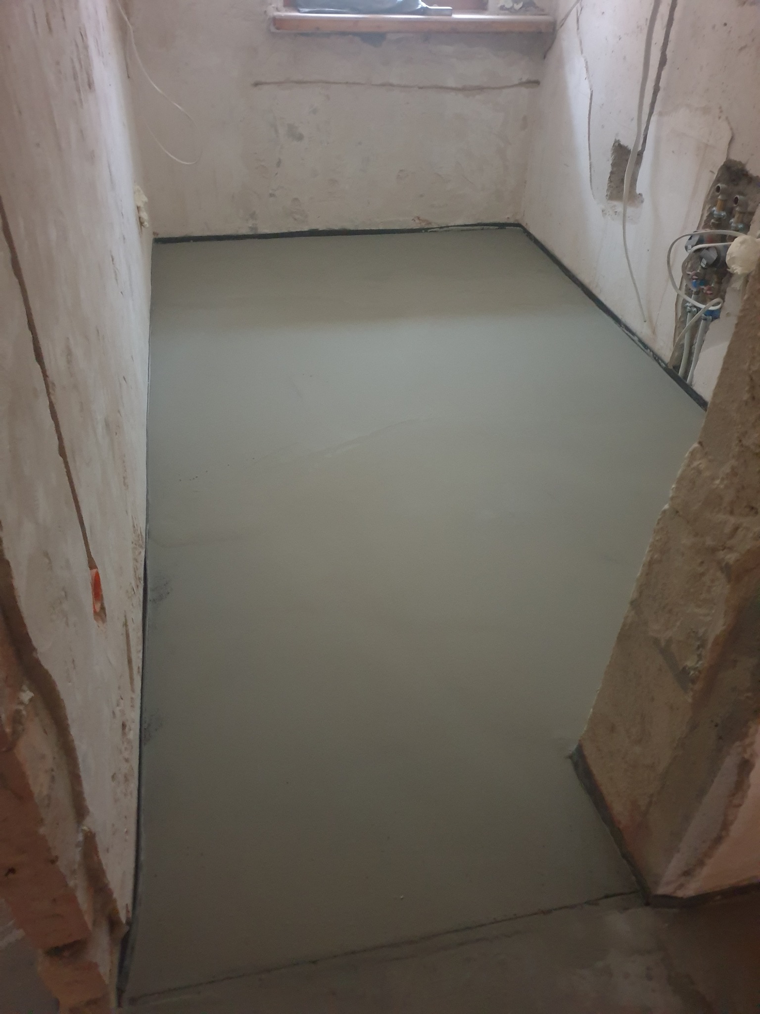 Wyrównana, świeża wylewka podłogowa w surowym wnętrzu. Widoczne fragmenty ścian w stanie deweloperskim i wystające instalacje. Minimalistyczny kadr, surowy klimat.