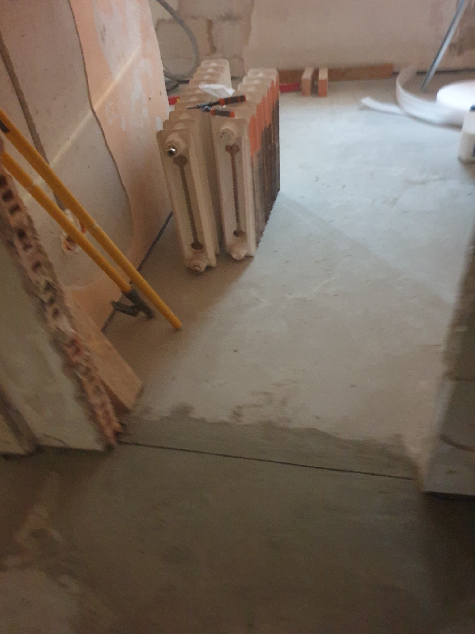 Wyrównana, szara posadzka w trakcie prac remontowych. W tle grzejnik żeberkowy, narzędzia i fragment ściany z widoczną warstwą izolacyjną. Ujęcie z niskiej perspektywy.