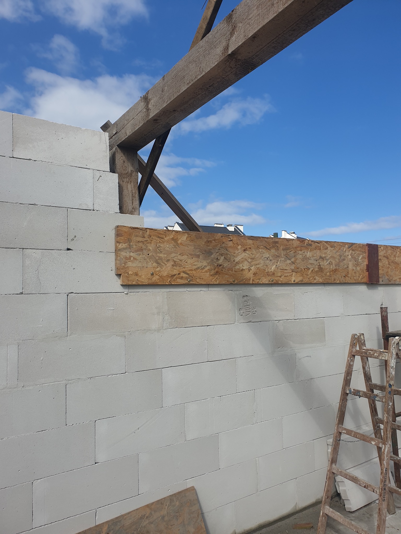 Ściana z białych bloczków, drewniana konstrukcja stropu i deska szalunkowa. Widoczne niebieskie niebo i drabina. Budowa domu w trakcie realizacji.