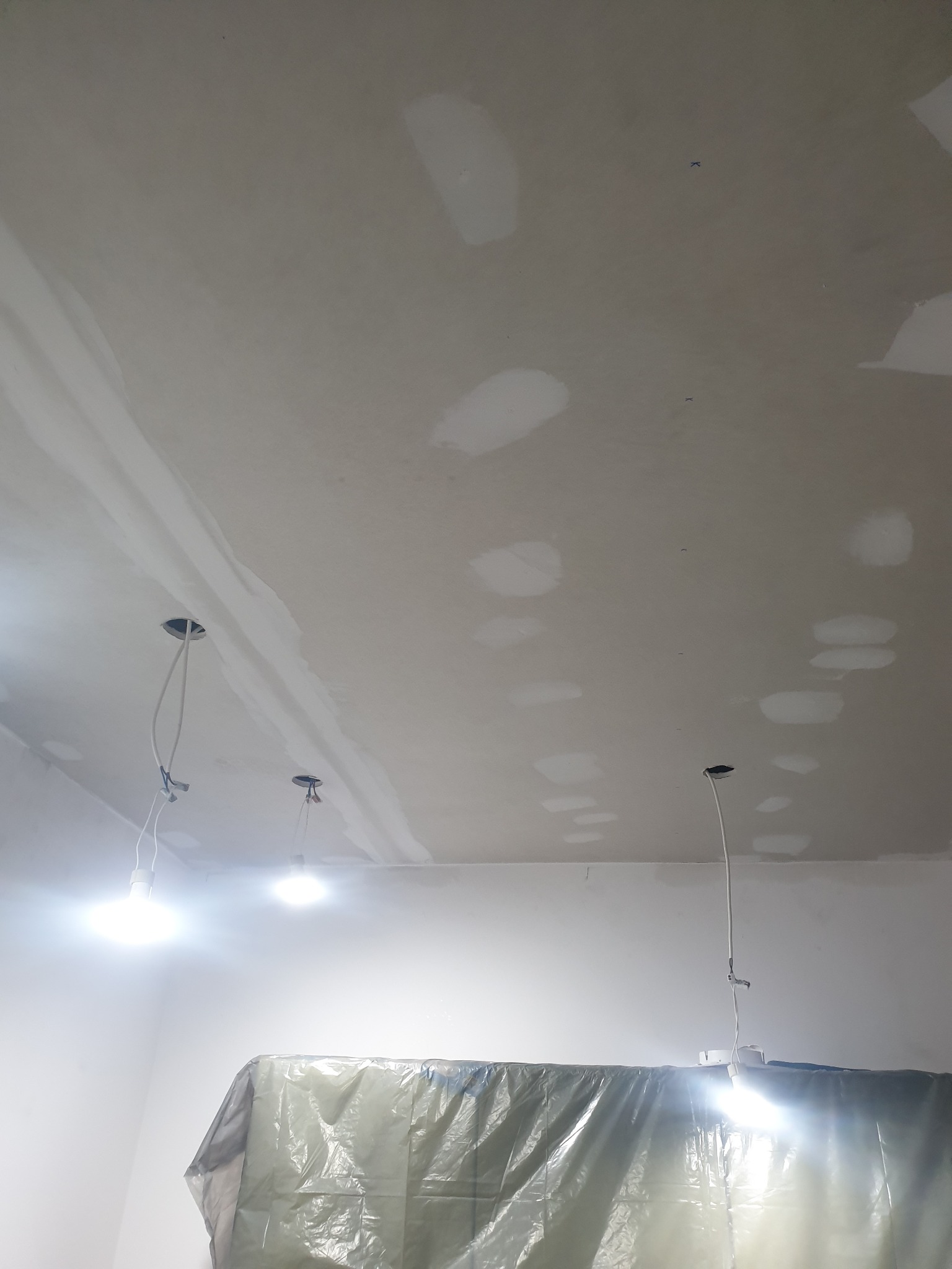 Wnętrze w trakcie prac wykończeniowych: sufit z widocznymi punktami świetlnymi i częściowo nałożoną gładzią, zabezpieczone folią meble. Widok z dołu.