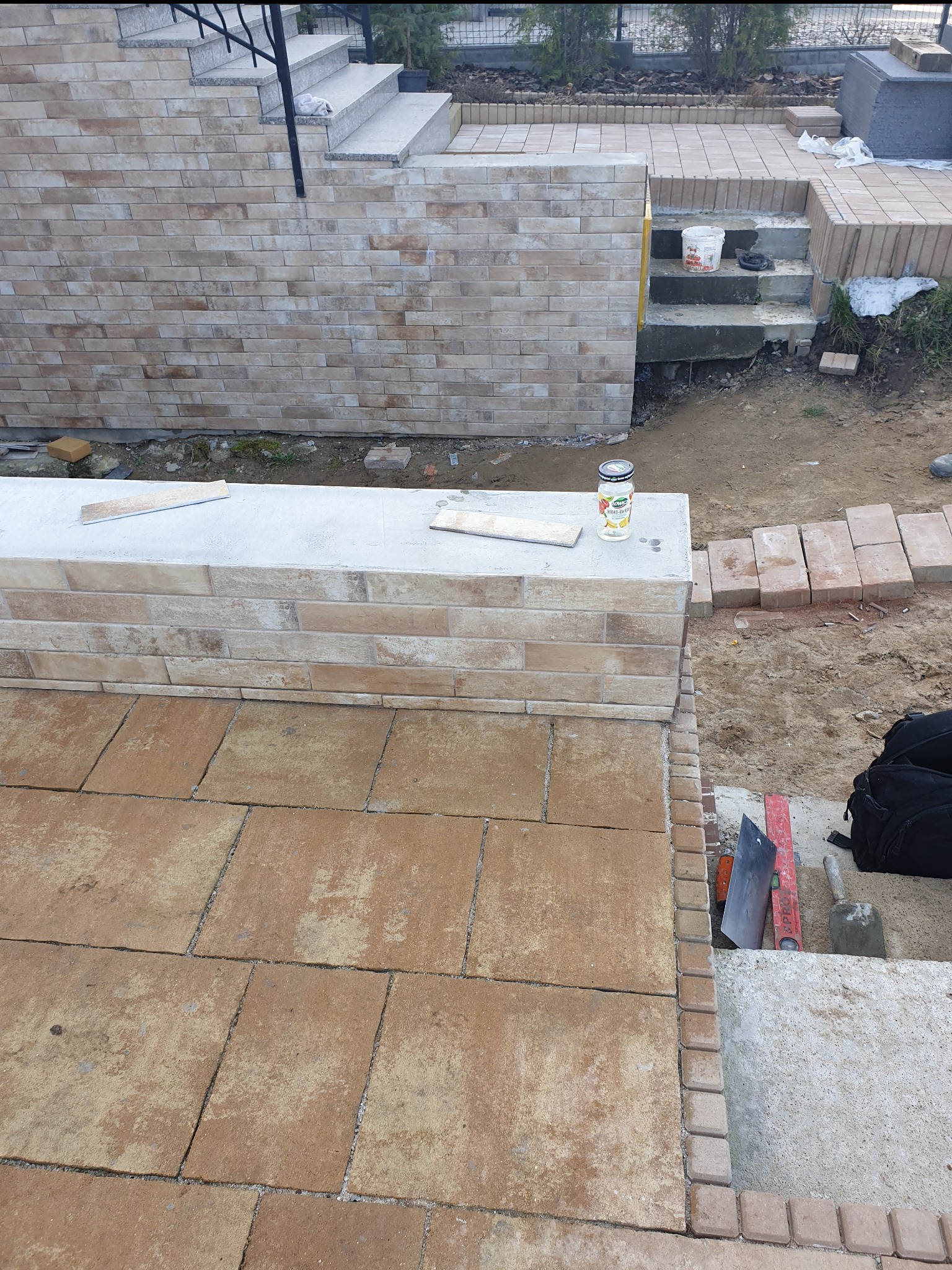 Układanie brązowej kostki brukowej na tarasie, widoczne narzędzia i fragment schodów z cegły. Prace w toku, zarys zieleni w tle.