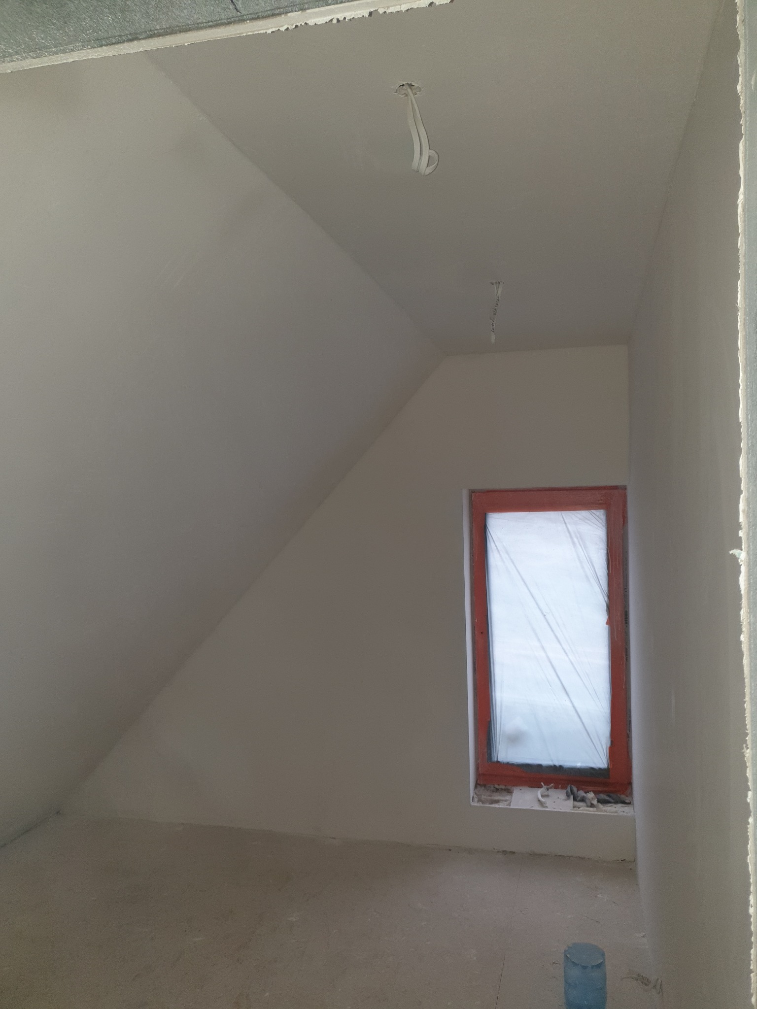 Poddasze po szpachlowaniu, okno zabezpieczone folią, widoczne kable oświetleniowe. Ściany i sufit gładkie, gotowe do dalszych prac wykończeniowych.