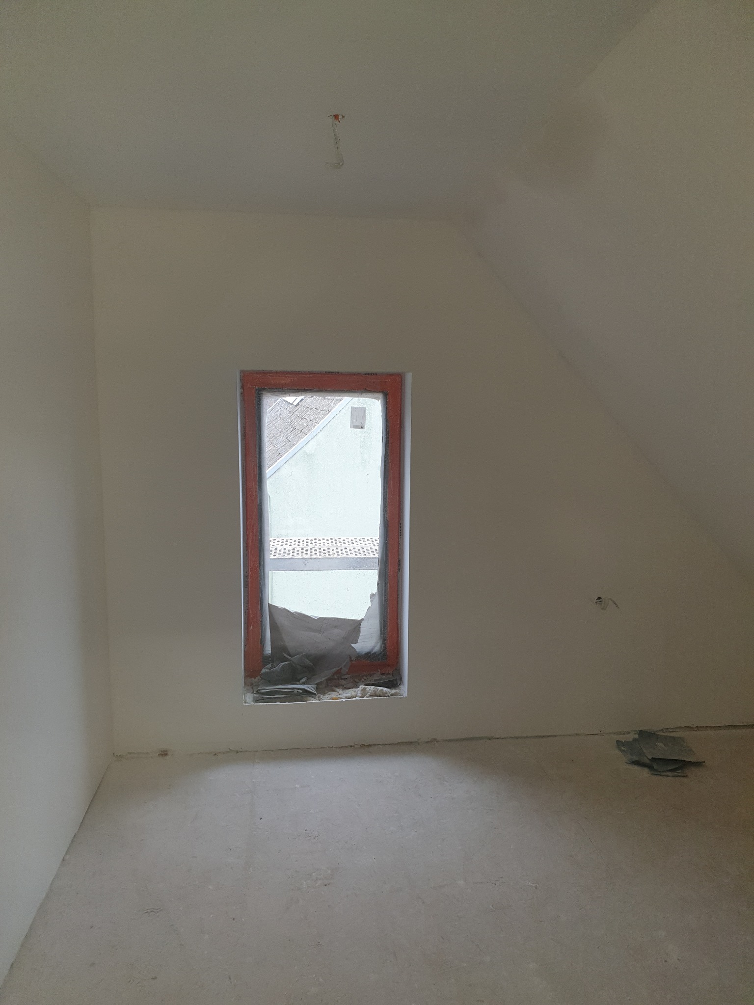 Poddasze po remoncie: gładkie, białe ściany, okno z pomarańczową ramą zabezpieczone folią. Widoczny fragment instalacji elektrycznej na suficie.