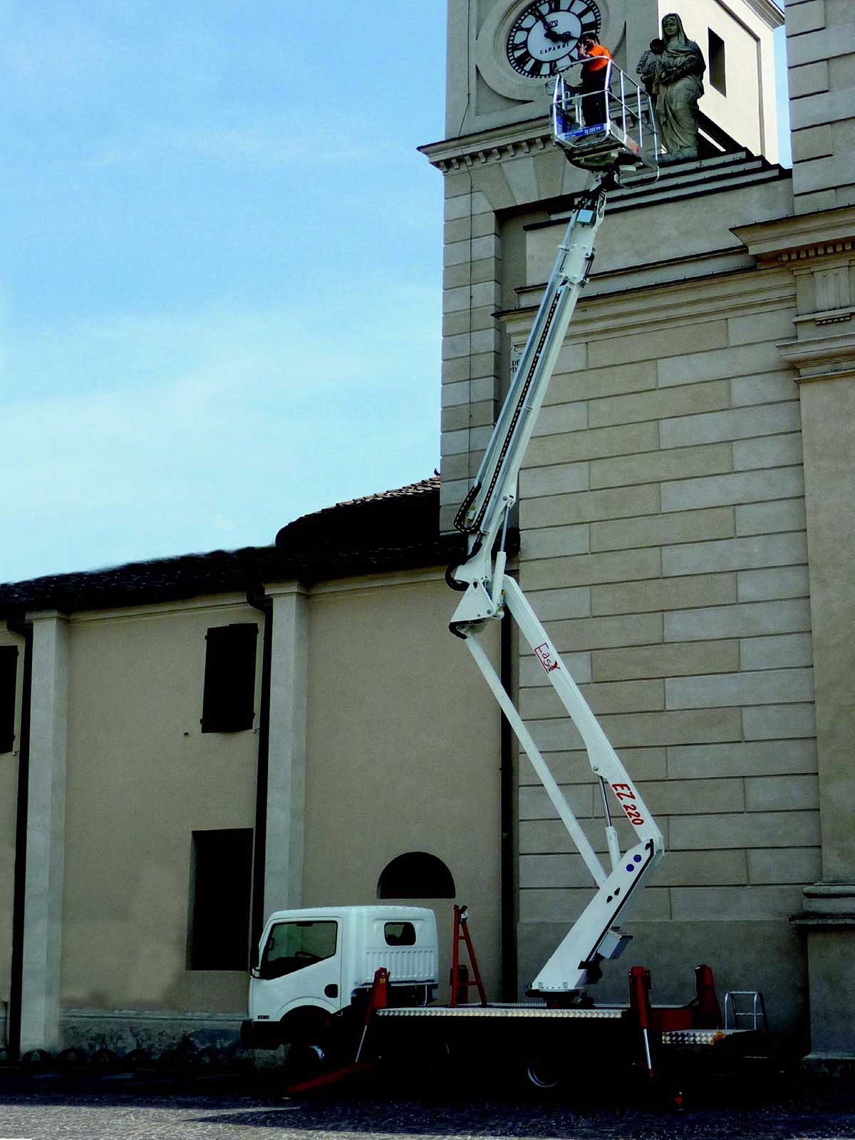 Biały podnośnik koszowy EZ 220 ustawiony przy fasadzie kościoła z zegarem i figurą Matki Boskiej, na platformie roboczej osoba w pomarańczowym kombinezonie.