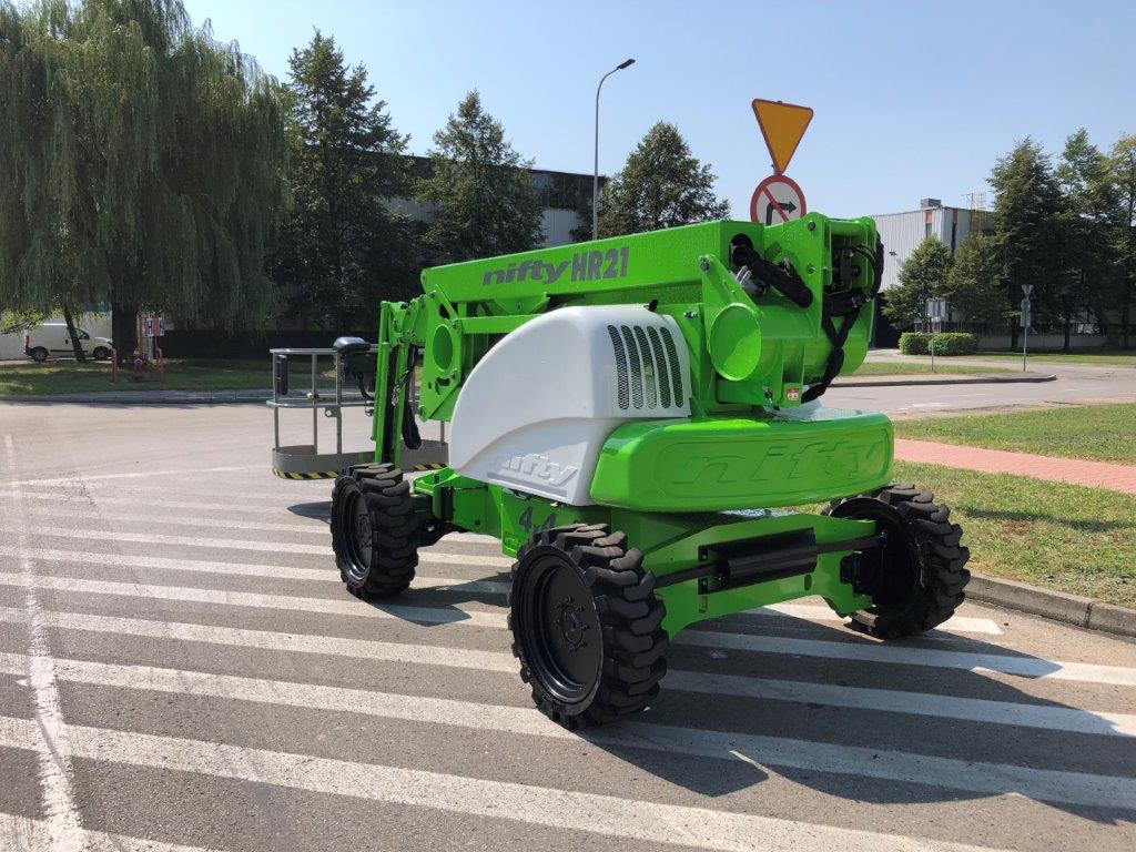 Zielony podnośnik koszowy Nifty HR21 na pasach dla pieszych, widok z boku, w tle budynek i drzewa.