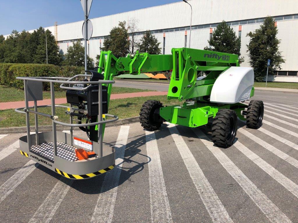 Zielony podnośnik koszowy Nifty HR21 na pasach dla pieszych, z widocznym koszem i logo niftylift.com, zaparkowany na tle budynku przemysłowego i zieleni.