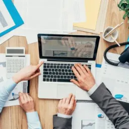 Widok z góry na biurko z laptopem wyświetlającym wykresy, kalkulatorem, telefonem stacjonarnym i dokumentami; dwie pary rąk wskazują na ekran laptopa i klawiaturę, sugerując analizę danych...