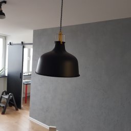 Usługi budowlane Piotr Gębski - Szara ściana z efektem dekoracyjnym, czarna lampa wisząca z tekstylnym kablem, widoczny fragment drabinki i roślina w tle.