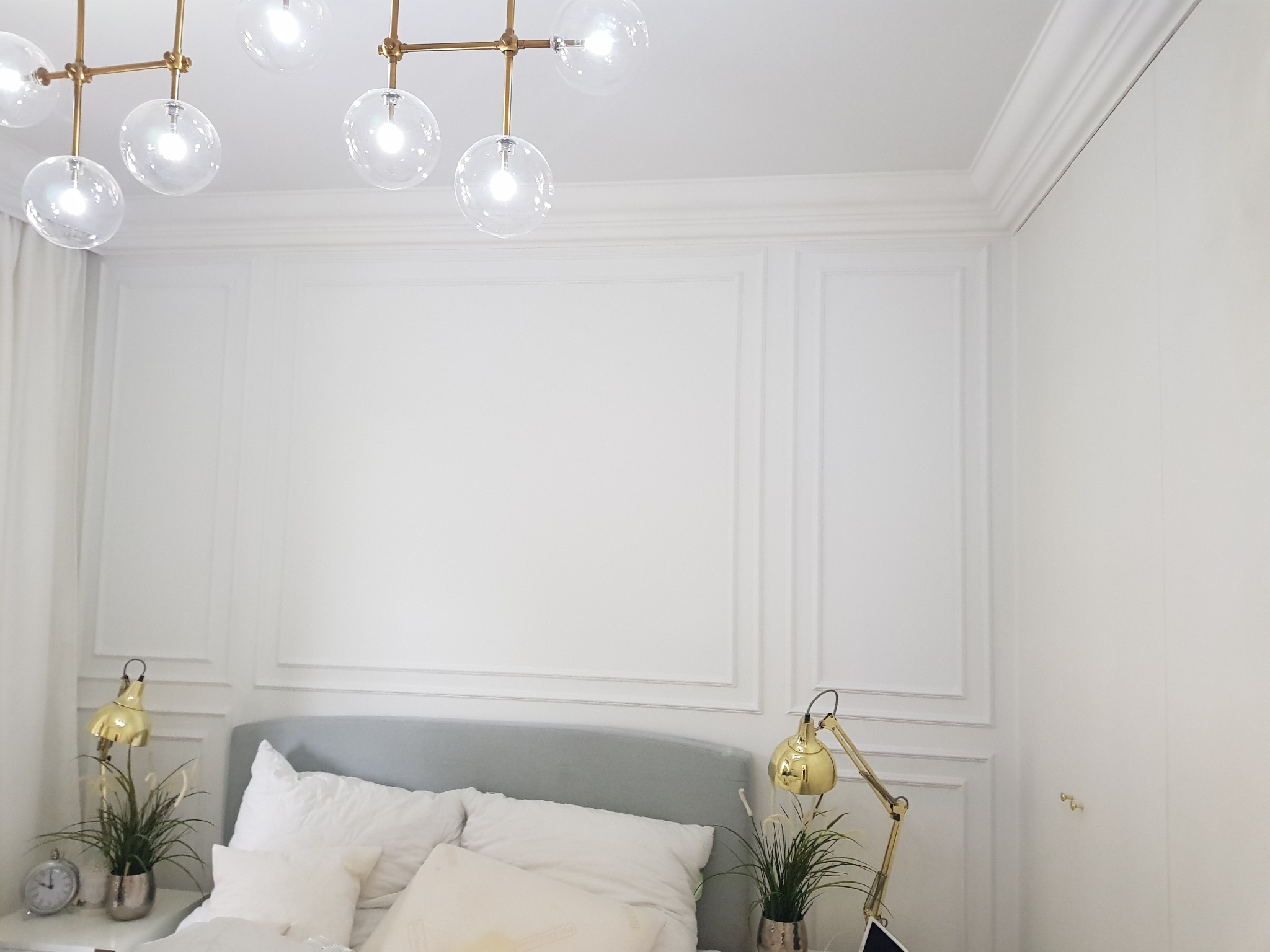 Elegancka sypialnia z białymi ścianami zdobionymi sztukaterią, szarą ramą łóżka i złotymi lampkami nocnymi, oświetlona nowoczesną lampą sufitową z kulistymi kloszami.