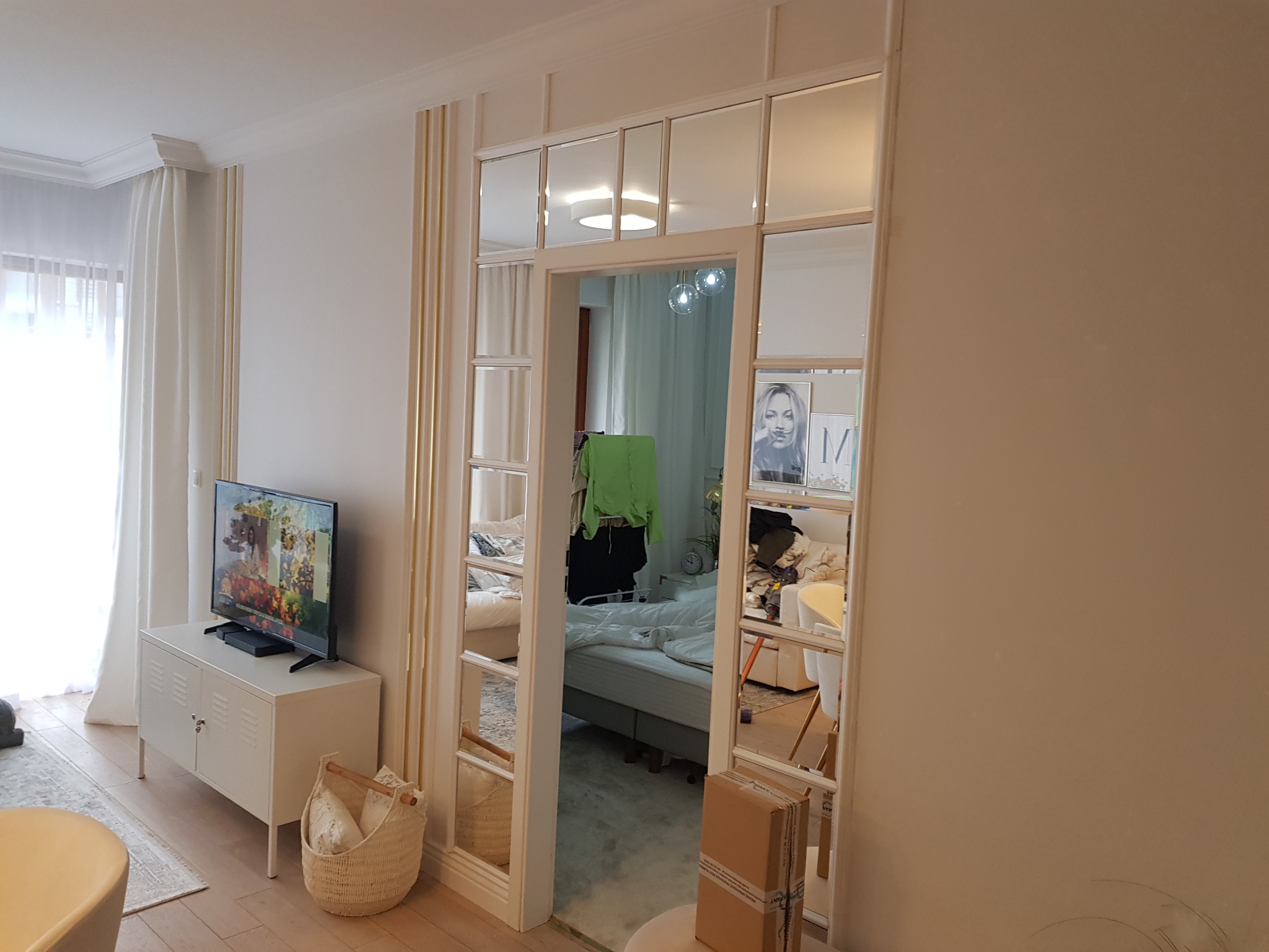 Salon z telewizorem i widokiem na sypialnię przez przeszklone drzwi z białymi ramami i złotymi akcentami.