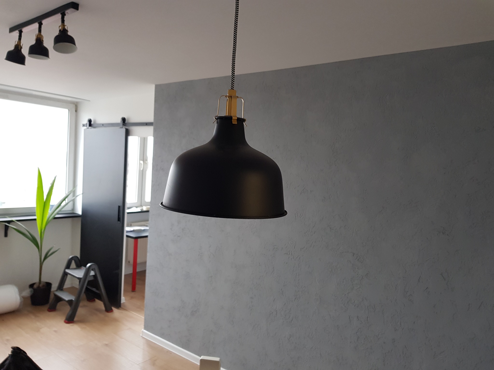 Szara ściana z efektem dekoracyjnym, czarna lampa wisząca z tekstylnym kablem, widoczny fragment drabinki i roślina w tle.