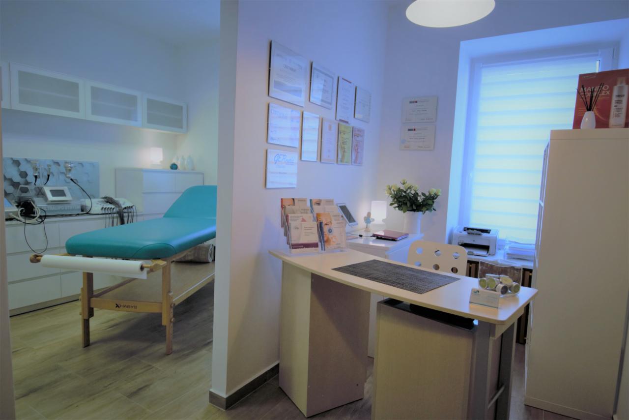 Jasne wnętrze gabinetu z turkusowym leżanką do zabiegów, biurkiem recepcyjnym z ulotkami i certyfikatami na ścianie, oświetlone lampą sufitową.