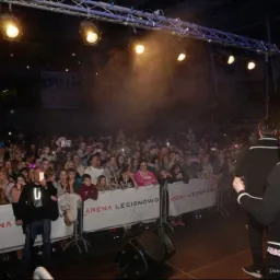 Koncert w Legionowie: tłum widzów przed sceną, oświetlenie punktowe skierowane na wykonawców, bariery z napisem ARENA LEGIONOWO oddzielające publiczność od sceny.