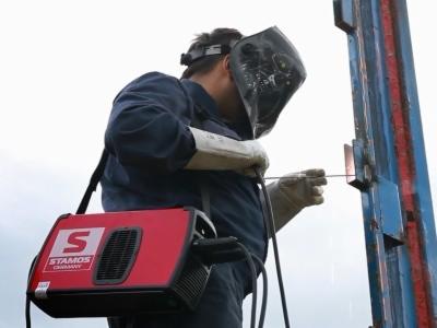 Pracownik w masce spawalniczej wykonuje spawanie elementu konstrukcji stalowej, z widocznym urządzeniem spawalniczym marki Stamos zawieszonym na ramieniu.