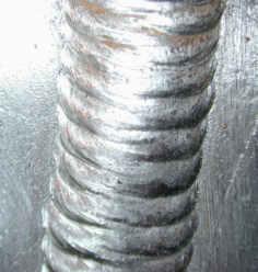 Szczegółowe zbliżenie spoiny pionowej wykonanej metodą spawania łukowego, widoczne regularne warstwy metalu o srebrzystoszarym odcieniu na surowej powierzchni metalu.