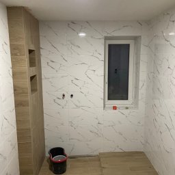 Bau Stone Kacper okoniewski - Wykończona łazienka w Nekli z białymi płytkami imitującymi marmur na ścianach i drewnopodobnymi płytkami na podłodze, widoczny fragment zabudowy z niszami i czarne wiadro.