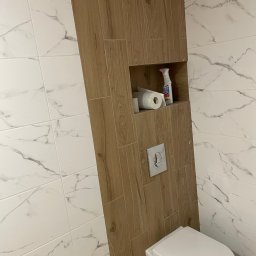 Bau Stone Kacper okoniewski - Wykończona toaleta z białą armaturą i drewnopodobną obudową, wbudowana półka z zapasem papieru toaletowego i środkiem czyszczącym, białe marmurkowe płytki na ścianach.