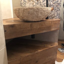 Bau Stone Kacper okoniewski - Kamienna umywalka nablatowa o nieregularnej fakturze, zamontowana na drewnianej szafce z półką, z czarną baterią, na tle mozaiki.