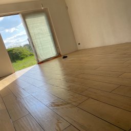 Bau Stone Kacper okoniewski - Ułożona podłoga z płytek imitujących drewno w jasnym, pustym pokoju z widokiem na zielony krajobraz przez otwarte drzwi balkonowe, częściowo zasłonięte folią ochronną.