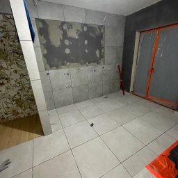 Bau Stone Kacper okoniewski - Wnętrze łazienki w trakcie układania płytek ceramicznych na ścianach i podłodze, widoczne przygotowane otwory na armaturę, fragment ściany z tapetą i drzwi zabezpieczone folią ochronną.