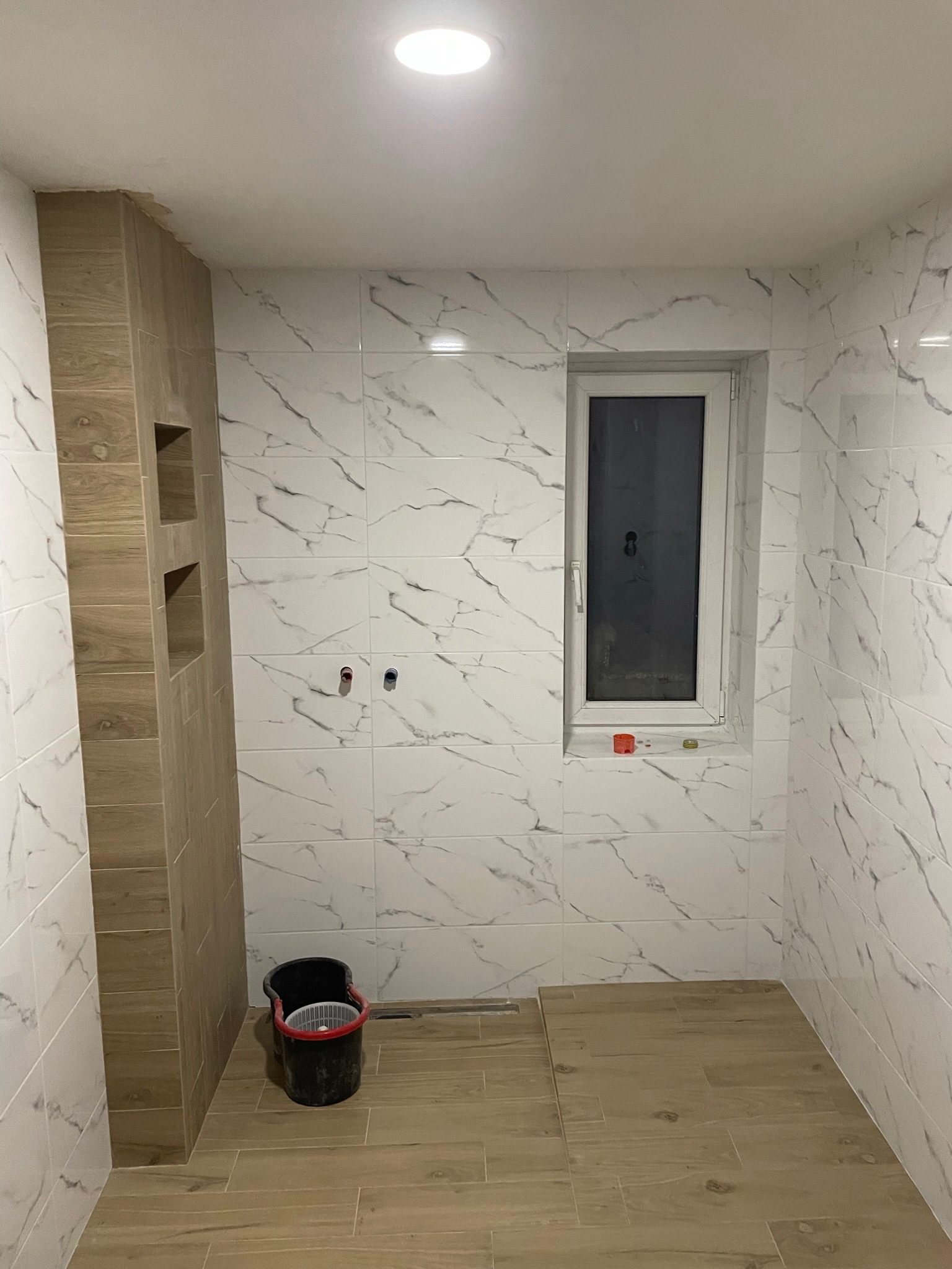 Wykończona łazienka w Nekli z białymi płytkami imitującymi marmur na ścianach i drewnopodobnymi płytkami na podłodze, widoczny fragment zabudowy z niszami i czarne wiadro.