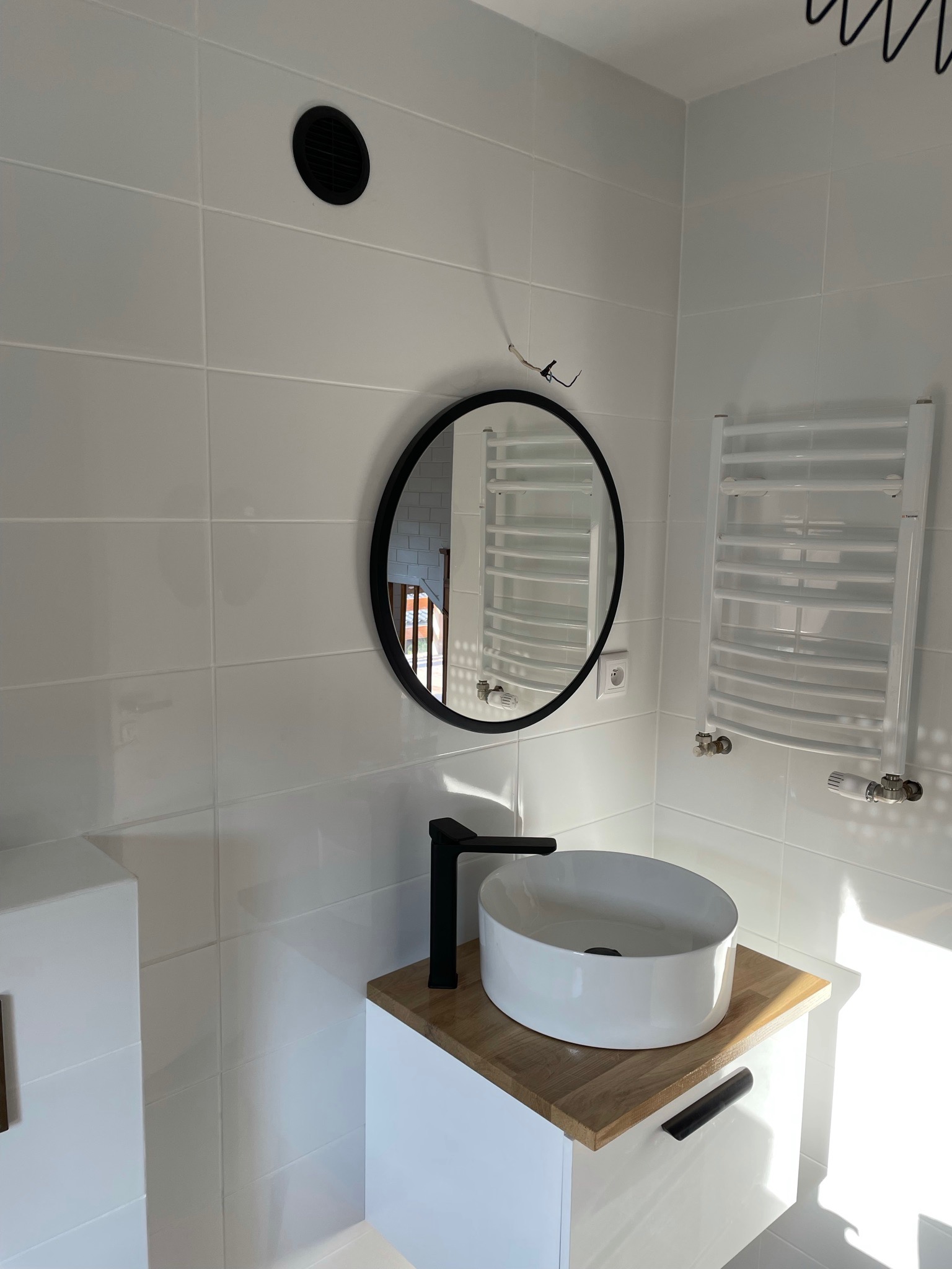 Nowoczesna łazienka z białą ceramiką, okrągłym lustrem w czarnej ramie, grzejnikiem drabinkowym i czarną armaturą na drewnianym blacie szafki pod umywalką.