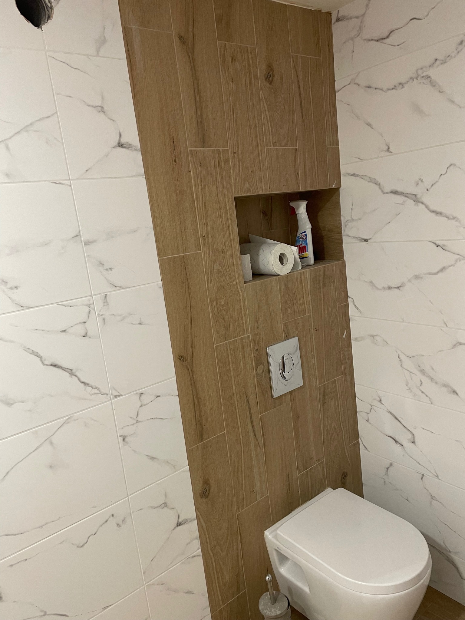 Wykończona toaleta z białą armaturą i drewnopodobną obudową, wbudowana półka z zapasem papieru toaletowego i środkiem czyszczącym, białe marmurkowe płytki na ścianach.