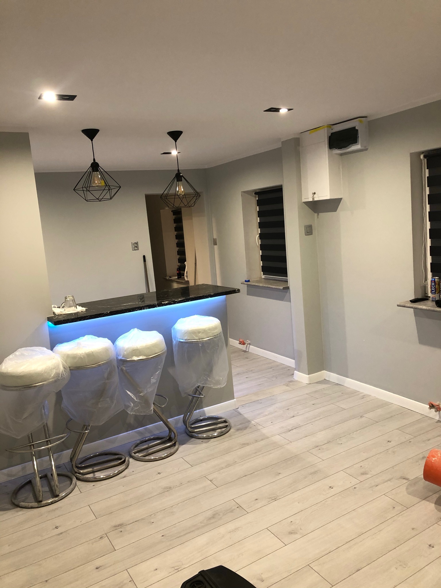 Nowoczesny bar z podświetleniem LED, krzesła barowe zabezpieczone folią, jasne panele podłogowe i geometryczne lampy wiszące w szarym wnętrzu po remoncie.