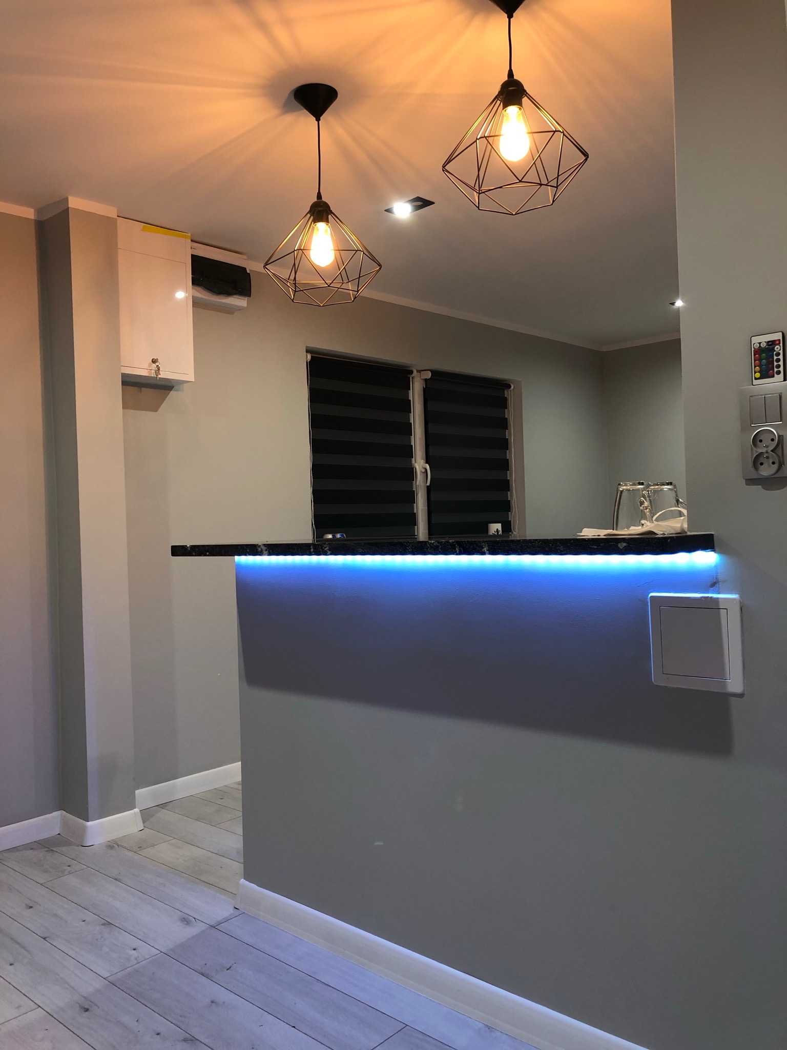 Nowoczesny aneks kuchenny z blatem podświetlonym niebieskimi diodami LED, geometryczne lampy wiszące, szare ściany i podłoga imitująca drewno.