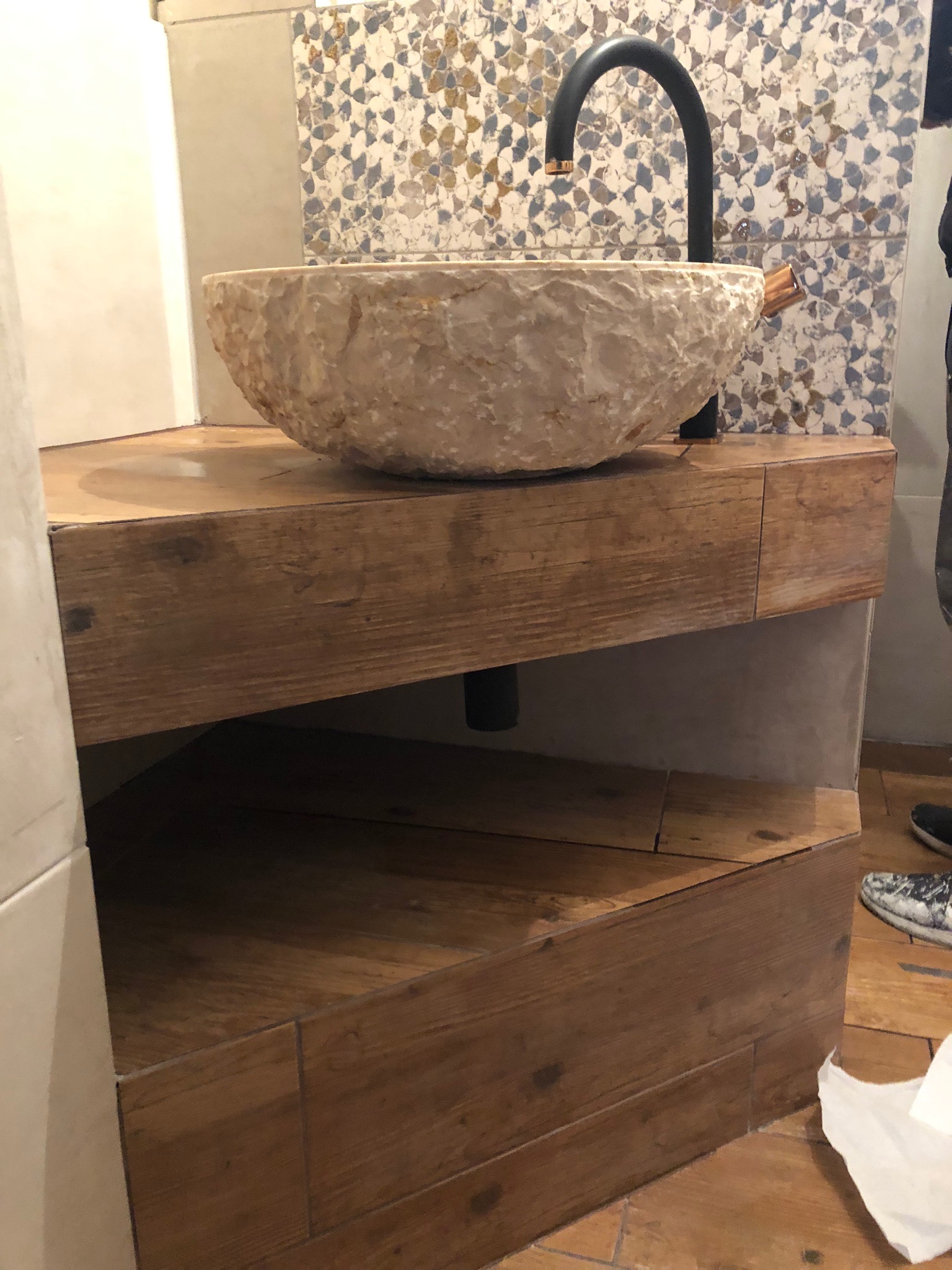 Kamienna umywalka nablatowa o nieregularnej fakturze, zamontowana na drewnianej szafce z półką, z czarną baterią, na tle mozaiki.