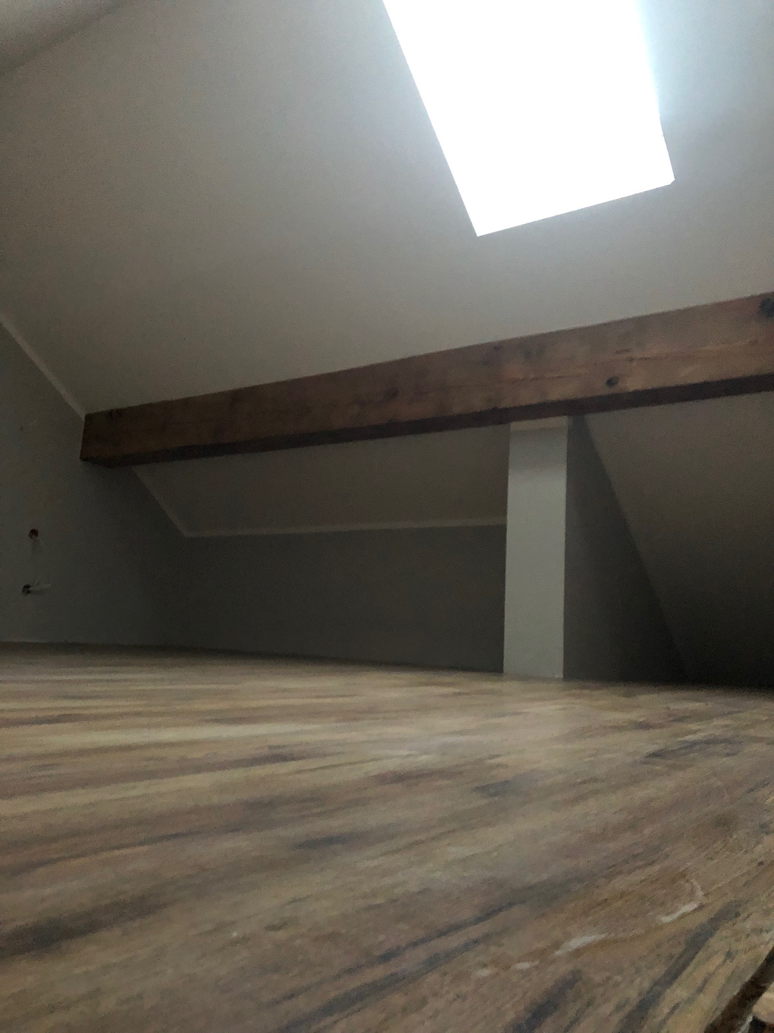 Wykończone poddasze z drewnianą podłogą, widoczna belka stropowa i okno dachowe w suficie. Ściany pomalowane na szaro.