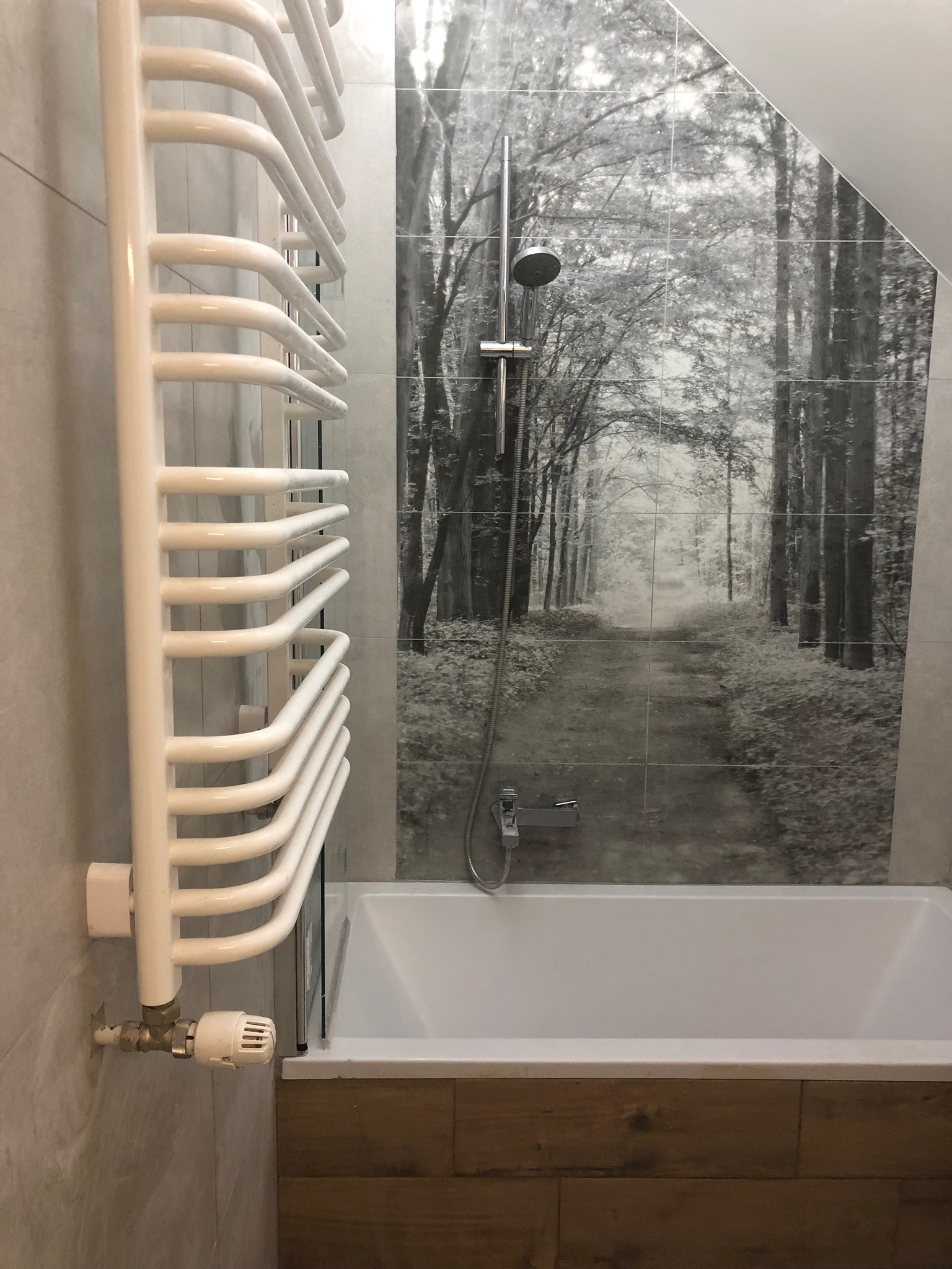 Biały grzejnik łazienkowy drabinkowy zamontowany na szarej płytce, widoczny termostat, w tle wanna obudowana drewnopodobnymi płytkami oraz ściana z płytek z motywem czarno-białego lasu...