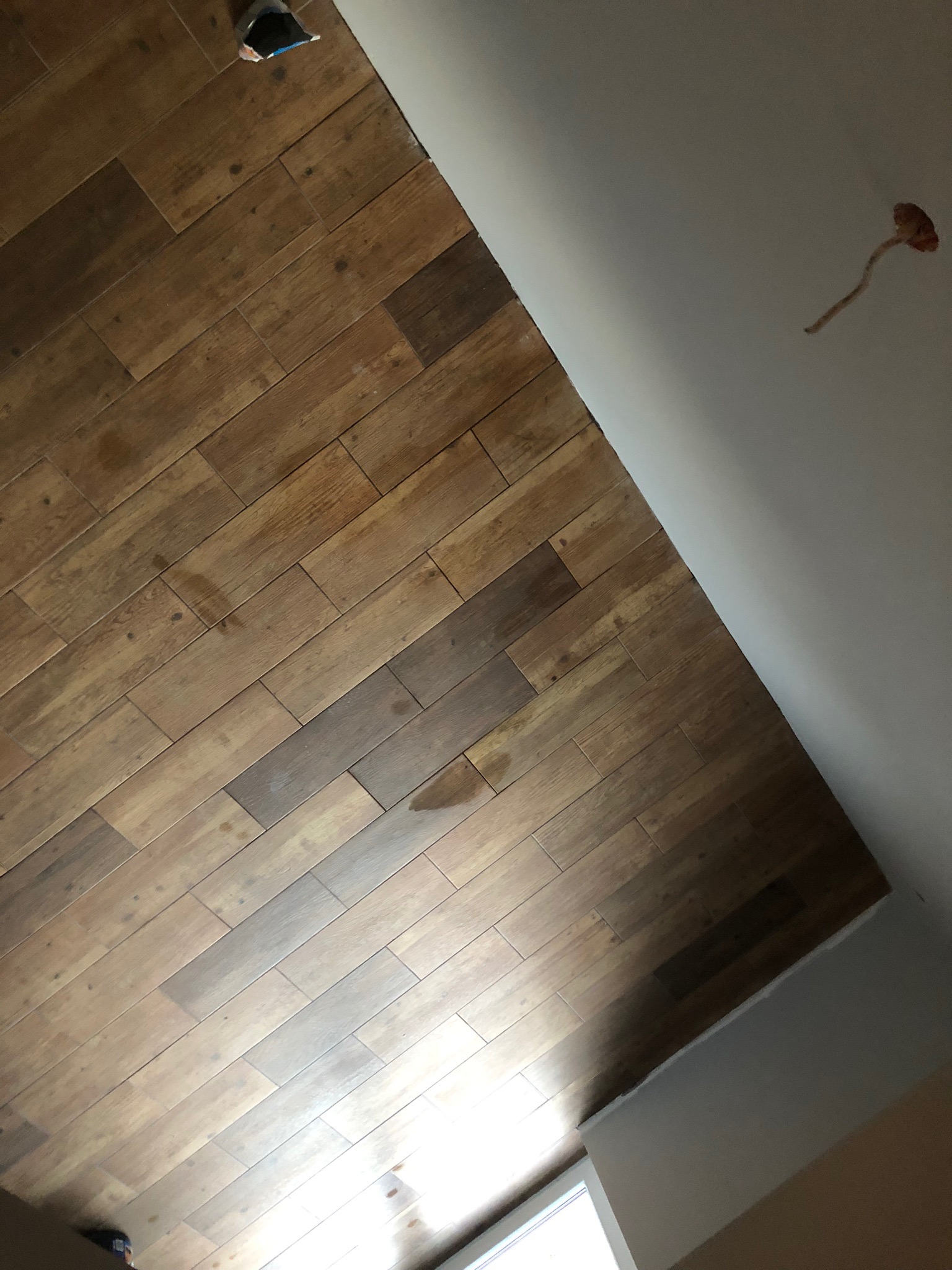 Ukośnie ułożona, drewnopodobna podłoga z płytek ceramicznych, widoczny fragment ściany z wystającą instalacją elektryczną i fragment okna w dolnym rogu.